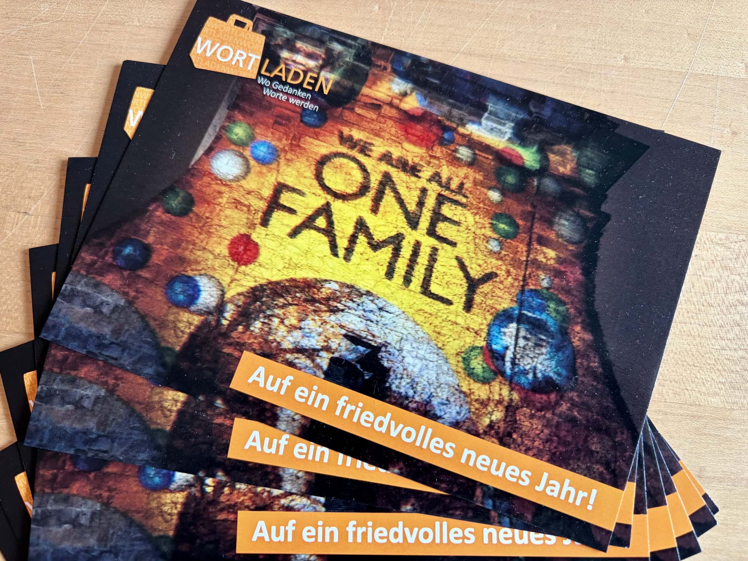 Foto: aufgefächerte Weihnachtskarten aus dem Wortladen. Ein friedvolles neues Jahr wird gewünscht, als Motiv ist eine beleuchtete Kirche zu sehen, auf der „We are all one Family“ projiziert wurde.